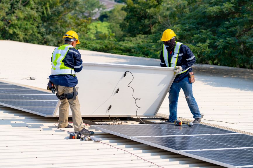 Two technicians in safety gear carefully removing a solar panel from a rooftop installation